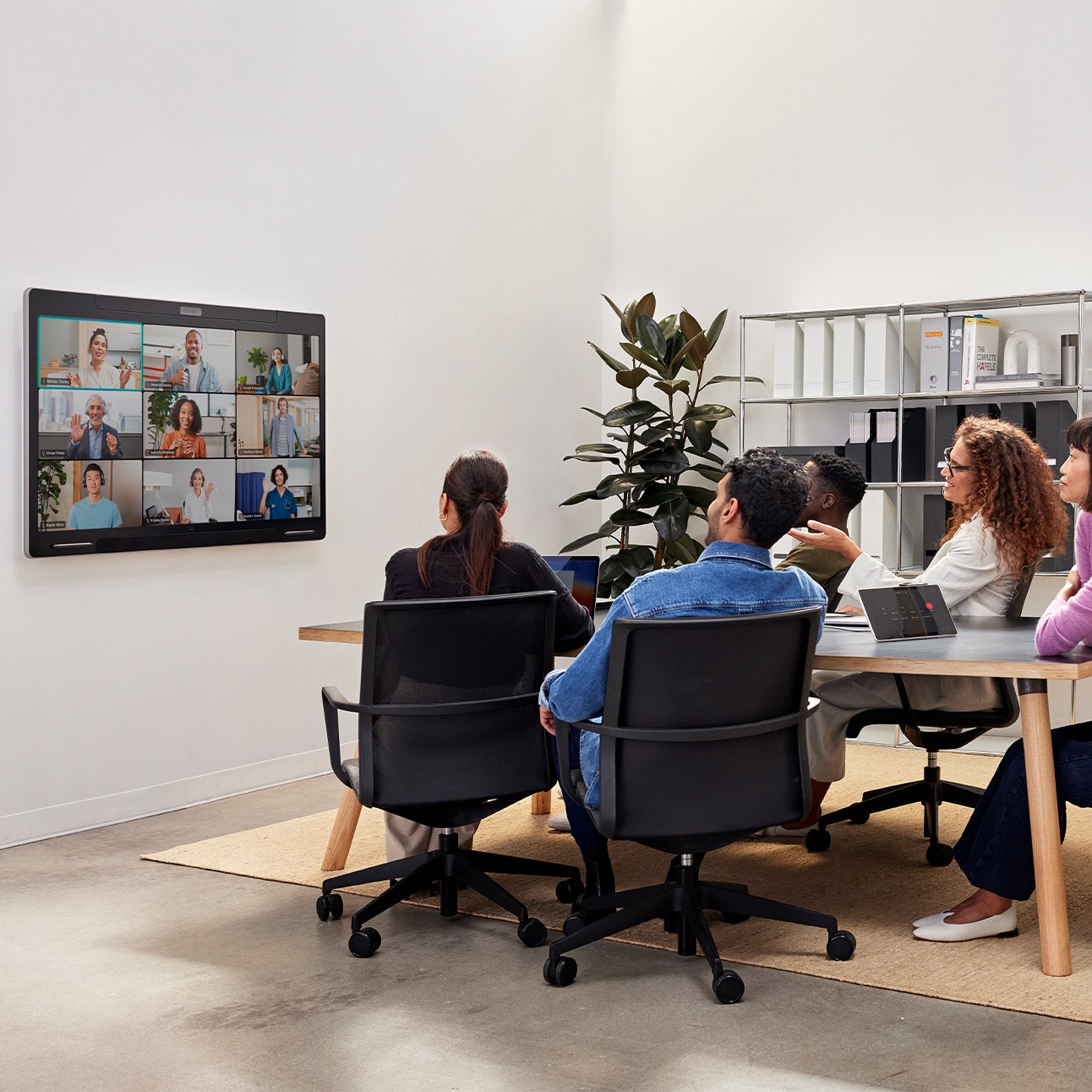 Five people collaborating in a small meeting room. Cisco Board Pro mounted on the wall showing remote participants in a grid view and the tabletop Cisco Room Navigator room controller.